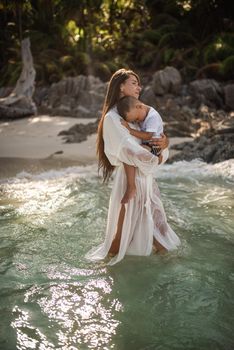 A young tall, long-haired brunette, a fair-haired girl, a mother, is walking along the azure sea and carries a small, fair-haired, happy son in her arms.white cotton clothes. boho dress.