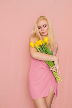 Adult blonde woman in pink dress posing over rosy background. Spring-summer clothing. She is holding bouquet of yellow tulips in her hands. Presents, surprise