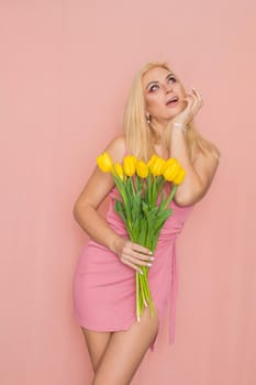 Adult blonde woman in pink dress posing over rosy background. Spring-summer clothing. She is holding bouquet of yellow tulips in her hands. Presents, surprise