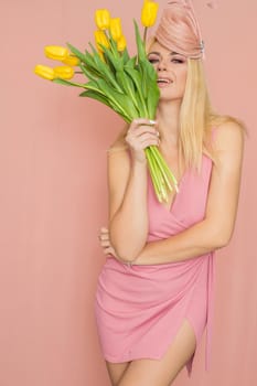 Adult blonde woman in pink dress posing over rosy background. Spring-summer clothing. She is holding bouquet of yellow tulips in her hands. Presents, surprise