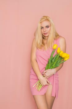 Adult blonde woman in pink dress posing over rosy background. Spring-summer clothing. She is holding bouquet of yellow tulips in her hands. Presents, surprise