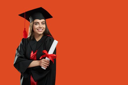 Front view of brunette girl standing with crossed hands, holding diploma. Pretty young woman graduating from college, university, high school. Isolated on red studio background.