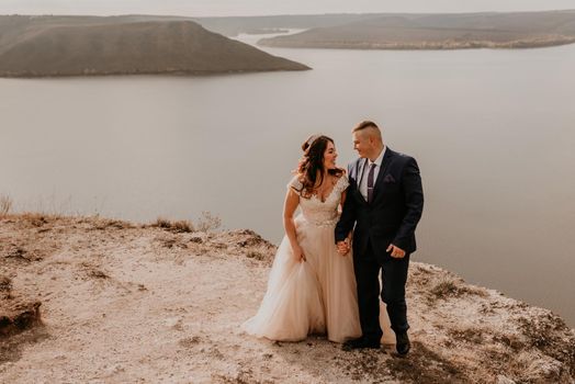 loving couple wedding newlyweds in white dress and suit walk in summer on mountain above river. sunset and sunrise. man and woman on rocks above cliff