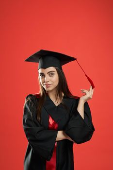 Front view of pretty girl in mortarboard and graduate gown standing with crossed hands. Pretty female student smiling, looking at camera. Isolated on red studio background,