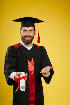 Front view of young male with beard graduating from college. Boy in graduate gown and mortarboard standing, showing diploma, smiling. Isolated on yellow studio background,