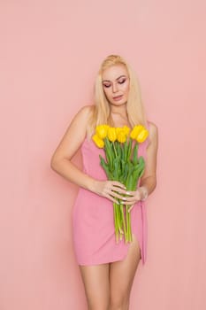 Adult blonde woman in pink dress posing over rosy background. Spring-summer clothing. She is holding bouquet of yellow tulips in her hands. Presents, surprise