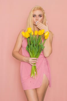 Adult blonde woman in pink dress posing over rosy background. Spring-summer clothing. She is holding bouquet of yellow tulips in her hands. Presents, surprise