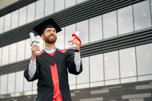 Front view of master graduating from college, university, high school. Male student in graduate gown and mortarboard standing, holding two diplomas, smiling. Concept of success.