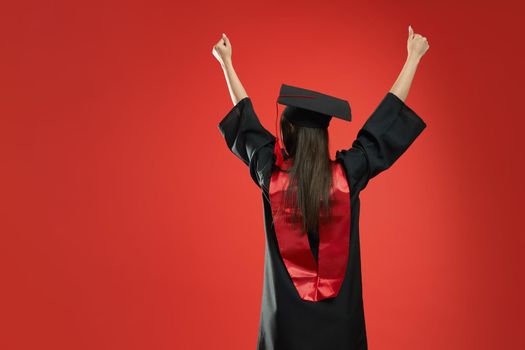 Back view of slim girl standing, graduating from college, university. Brunette young female with long hair showing hurray, wearing mortarboard and graduate gown. Concept of youth.