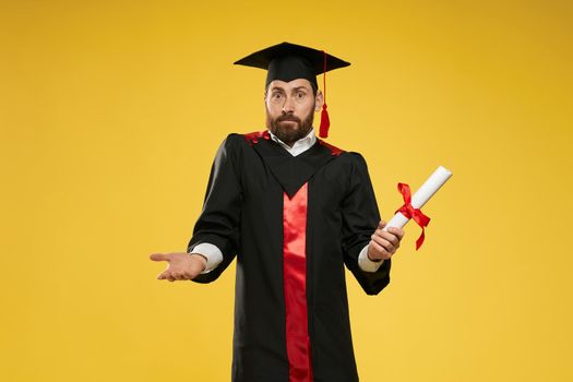 Front view of male with beard standing, holding diploma, surprised, mixed up. Handsome boy graduating from college, university, high school. Isolated on yellow studio background.