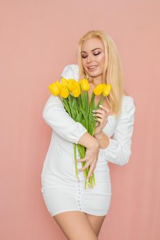 Spring fashion photo of blonde woman posing over pink background. Wearing white vintage dress with long sleeves. Holding bouquet with yellow tulips in her hands