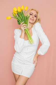 Spring fashion photo of blonde woman posing over pink background. Wearing white vintage dress with long sleeves. Holding bouquet with yellow tulips in her hands