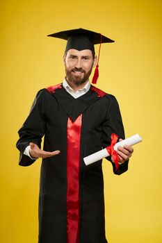 Front view of proud, cheerful student graduating from university. Young male with beard in graduate gown and mortarboard standing, holding, showing diploma, Concept of knowledge.