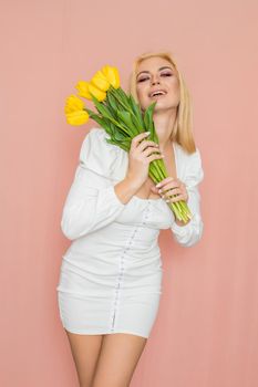Spring fashion photo of blonde woman posing over pink background. Wearing white vintage dress with long sleeves. Holding bouquet with yellow tulips in her hands