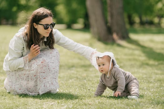 A young mother is playing with her 7-month daughter in the meadow. An infant girl is crawling on the grass.