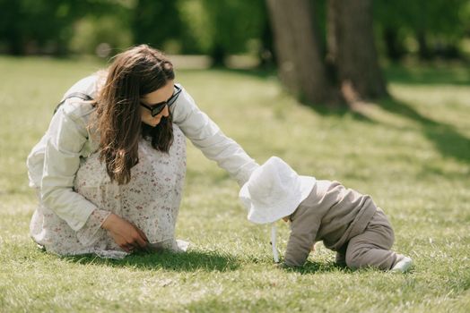 A young mother is playing with her 7-month daughter in the meadow. An infant girl is crawling on the grass.