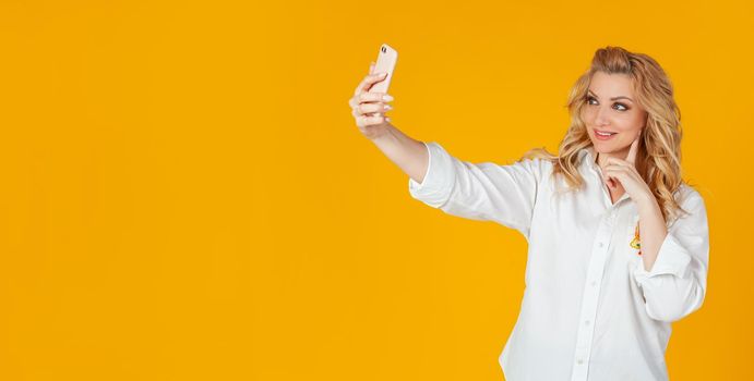 Portrait of a stylish beautiful smiling caucasian blonde woman taking a selfie on a smartphone, holding a mobile phone in outstretched hand and trying to look beautiful in the photo, yellow background