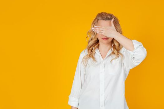 Portrait of a woman closes her eyes with the palm of her hand. Blonde waiting for an unexpected gift, waiting for the present, counting ten, playing hide and seek, standing on a yellow background