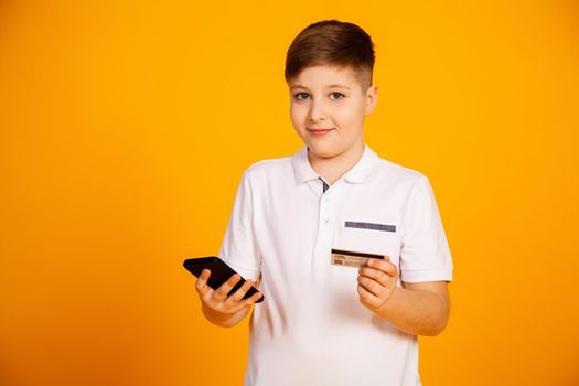 The boy holds a credit card and the smartphone makes purchases. Happy young man uses a smartphone and a bank card to shop online. Handsome smiling guy holds a bank card and mobile phone