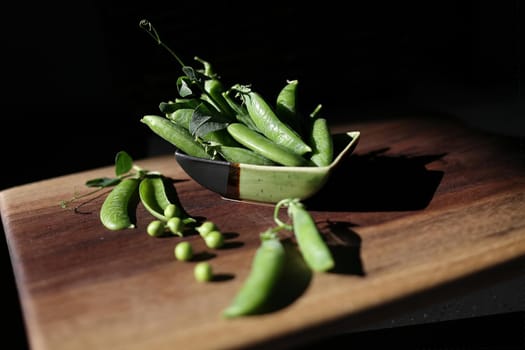 Delicious ripe green peas lie in a small ceramic bowl and on a wooden table. Dark background, rustic style, hard sunlight, selective focus