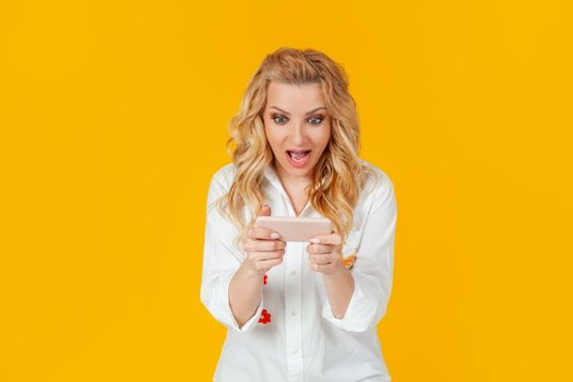 Cheerful excited pretty woman plays a new amazing game for smartphones, screams joyfully and smiles, winning, on a yellow background