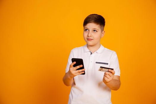 The boy holds a credit card and the smartphone makes purchases. Happy young man uses a smartphone and a bank card to shop online. Handsome smiling guy holds a bank card and mobile phone