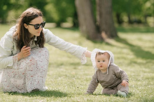 A young mother is playing with her 7-month daughter in the meadow. An infant girl is crawling on the grass.