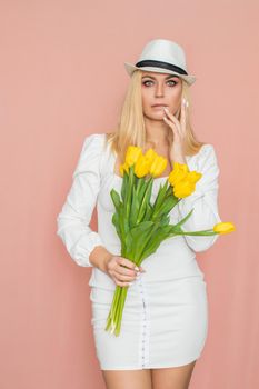 Spring fashion photo of blonde woman posing over pink background. Wearing white vintage dress with long sleeves and white hat. Holding bouquet with yellow tulips in her hands