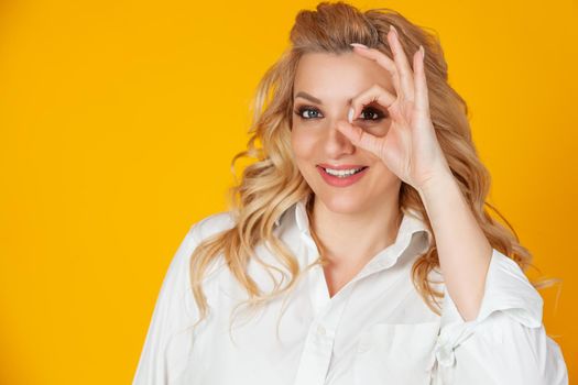 Blond woman feeling optimistic happy showing ok circle over eye smiling happily. feels happy optimistic showing ok, good gesture okay with an eye sign smiling happily standing yellow background