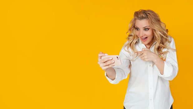 Technology, online lifestyle concept. Caucasian blond woman rejoicing in victory in the game, showing her finger on a smartphone, saying yes, cheers, pleased with a smile on a mobile phone,