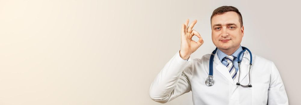 Good job. Satisfied doctor man in a medical coat and with a stethoscope shows okay okay sign of approval smiles, white background. No problem, clinic guarantee best professional service and caring.