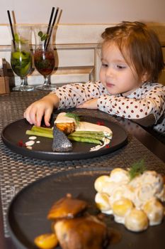 A little girl in a restaurant looks at a freshly brought fish dish on the table in front of her and wants to take some caviar from the plate with her hands to taste it. the dish looks very appetizing and the daughter could not resist and tries it right with her hands. in the foreground in the frame potatoes in mushroom sauce and pieces of meat
