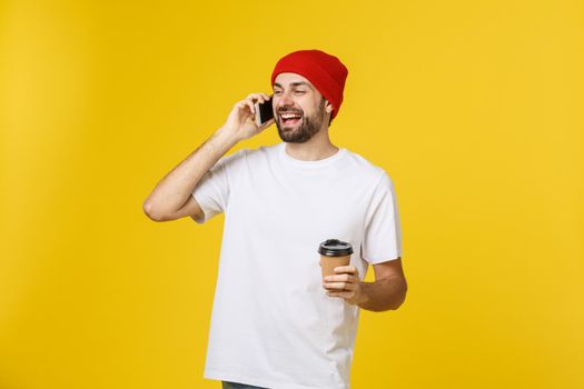 Portrait of a cheerful young man wearing casual clothes standing isolated over yellow background, holding mobile phone, drinking takeaway coffee