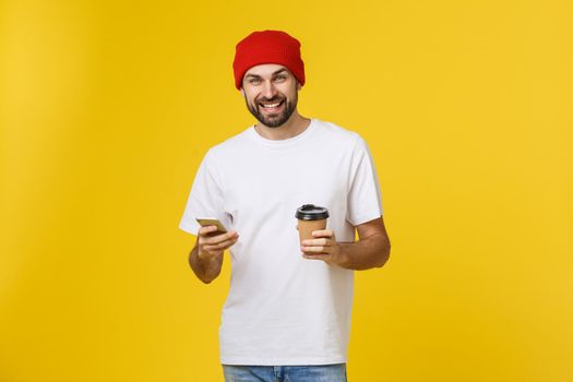 Portrait of a cheerful young man wearing casual clothes standing isolated over yellow background, holding mobile phone, drinking takeaway coffee