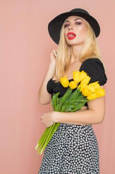 Summer fashion portrait blonde woman. Sexy look in black top and skirt, wearing hat. Red lips. Holding yellow tulips in her hands