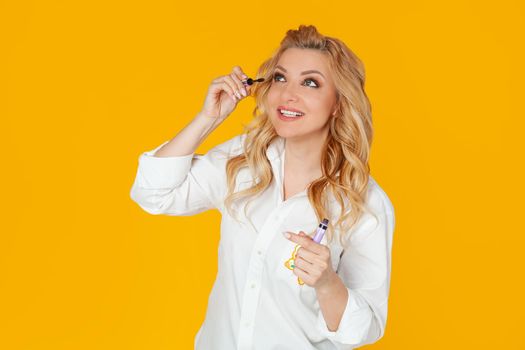 Blonde in white shirt, middle aged European woman smiles broadly, holding and applying mascara, standing against yellow background