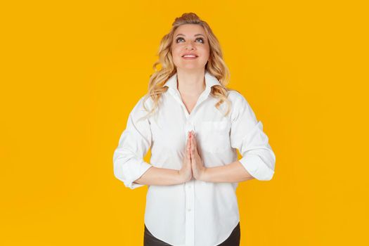 Cheerful attractive happy European blonde woman in white shirt, holding hands to her chest, making a wish, smiling, raising her head to the sky, waiting for good news, praying. yellow background