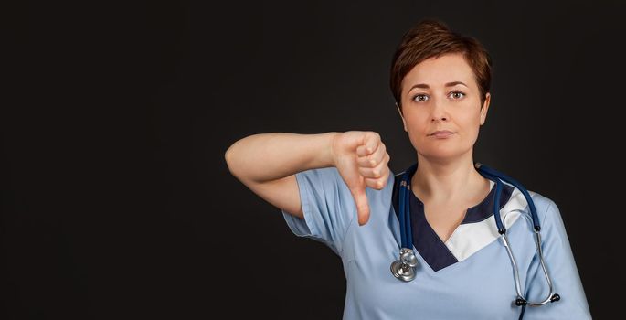 Discontent European doctor woman shows disapproval sign, keeps thumb down, expresses dislike, frowns face in discontent, isolated over black background. Body language concept.