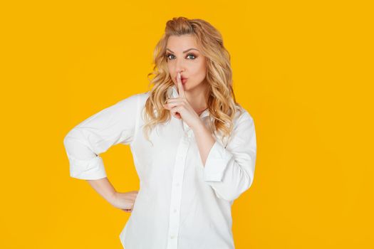 A domineering, serious European blonde woman in a white shirt, says shut up, be quiet, shielding the camera, pressing her index finger to her lips, scolding for being too loud.