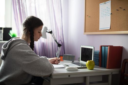 At home in her room, the teenager sits at her desk and solves assignments from school. A diligent student. Remote learning at home.