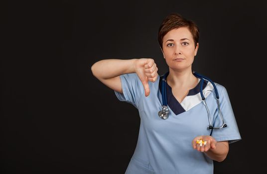 Discontent European doctor woman shows disapproval sign, keeps thumb down, expresses dislike, frowns face in discontent, isolated over black background. Body language concept.