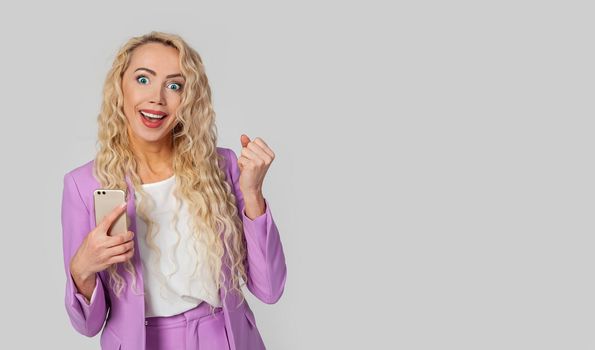 A happy triumphant woman holds a smartphone, fist, and an enthusiastic smile receives great fantastic news using a text message on her mobile phone. Wins the online lottery, receives the first prize.