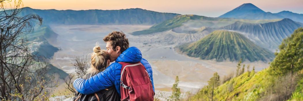 BANNER, LONG FORMAT Young couple man and woman meet the sunrise at the Bromo Tengger Semeru National Park on the Java Island, Indonesia. They enjoy magnificent view on the Bromo or Gunung Bromo on Indonesian, Semeru and other volcanoes located inside of the Sea of Sand within the Tengger Caldera. One of the most famous volcanic objects in the world. Travel to Indonesia concept.