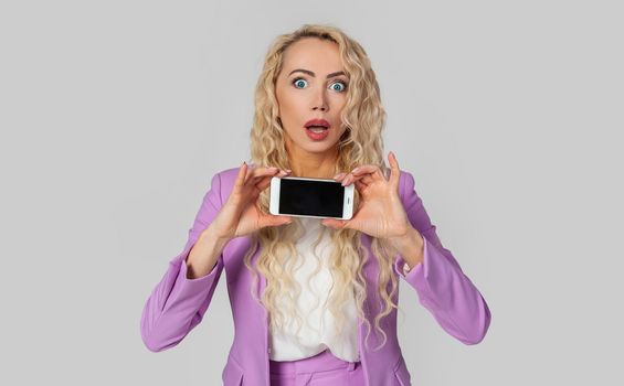 Portrait of amazed blonde woman impressively showing smartphone screen, holding mobile phone horizontally, amusing open mouth