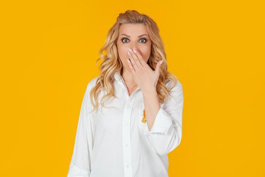 Blandin european middle-aged woman in white shirt, panic in shock upset, frowning, covering her mouth with her hand, disappointed to hear bad news unhappy standing on a yellow background.