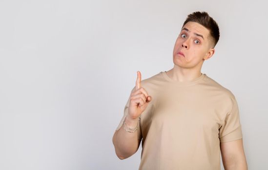 A handsome guy with a beautiful haircut looks at the camera with a funny facial expression, holding his lips and pointing his finger up. Copy space