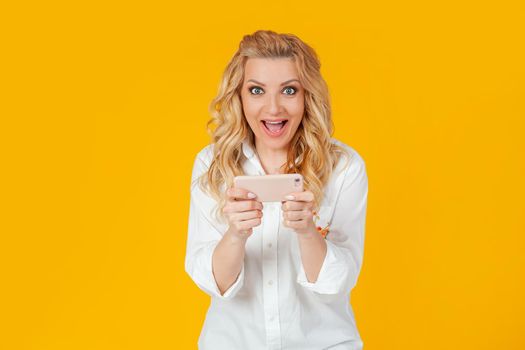 Excited European blonde woman in white shirt, looks surprised and excited, plays awesome new smartphone game, screams and smiles happy, wins. Standing on a yellow background