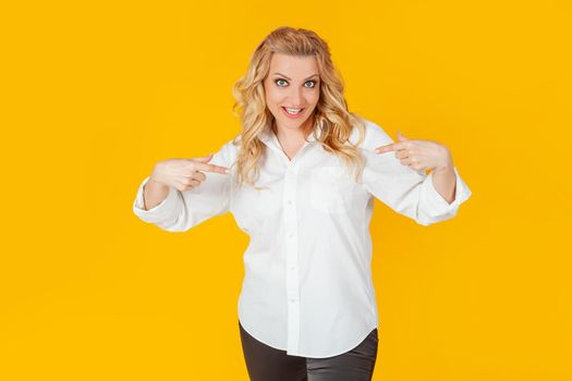 Amazed speechless impressed happy European blonde woman in white shirt pointing at herself shocked, with open mouth, drooping jaw, amazed, to receive an unexpected great offer on a yellow background.
