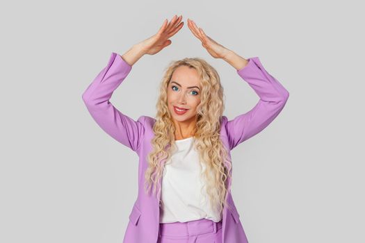 Life insurance policy. Happy adult woman with curly hair in a business suit making a gesture of a house roof over her head with her hands, feeling safe and protected, dreaming of buying a house.