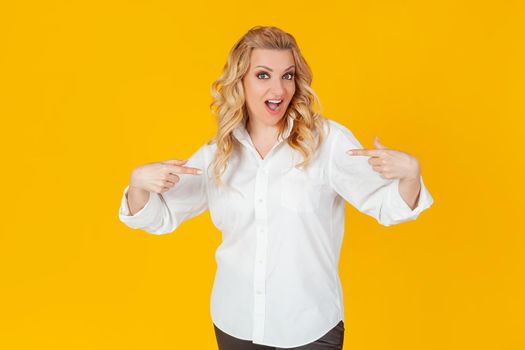 Amazed speechless impressed happy European blonde woman in white shirt pointing at herself shocked, with open mouth, drooping jaw, amazed, to receive an unexpected great offer on a yellow background.